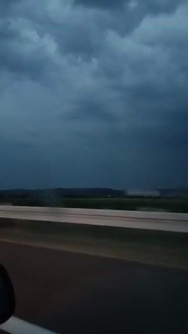 The video shows a view from a moving vehicle looking out at a dark, stormy sky over a rural landscape. The sky is filled with heavy, dark clouds, suggesting an impending storm. In the distance, there are rolling hills or fields, with some patches of green vegetation visible. A highway or road is in the foreground, with a white lane divider. The overall lighting is dim due to the overcast conditions. The camera appears to be mounted inside the vehicle, with a portion of the vehicle's interior visible in the bottom left corner. There are no people visible in the frame, and no specific actions are taking place other than the movement of the vehicle.