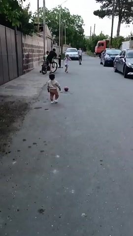 The video shows a street scene with several children and adults. In the foreground, a young child in a light brown outfit stands facing the camera, with a red ball to their left. Further up the street, a young girl in a white dress is running towards the camera. To the left, a person on a bicycle is partially visible. In the background, two other children are on scooters, and an adult is standing and observing. There are cars parked along the right side of the street, and buildings and trees line the left and background. The overall scene appears to be a casual outdoor gathering or playtime.