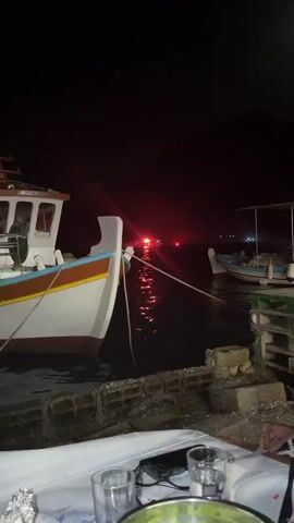 The video shows a nighttime scene at a harbor or marina. In the foreground, the side of a white boat with blue and brown trim is visible, tethered by a rope. The boat is docked next to a stone wall. In the background, across the dark water, another boat is seen with its lights on. This boat has multiple red flashing lights and appears to be emitting smoke or steam, suggesting a possible incident or activity such as a distress signal or a fire suppression effort. The red lights reflect on the surface of the water. Another smaller, less distinct light can be seen further in the distance.