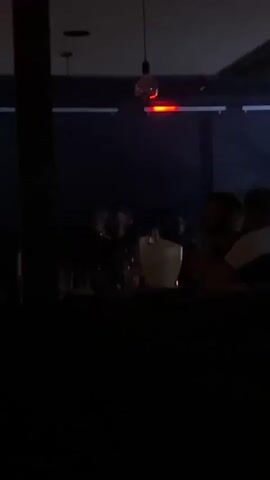 Here's a brief description of the video:

The video shows a dimly lit bar with several people sitting at the counter. One man is seen pouring a bottle into an ice bucket. Red and blue lighting shifts in the background, adding to the ambiance of the bar. A lightbulb hangs from the ceiling. The focus is on the actions at the bar and the people interacting there.