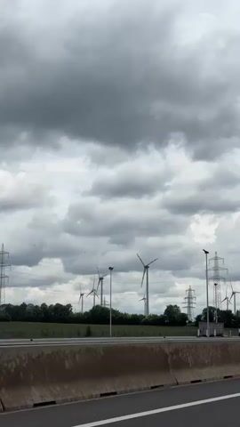 From the perspective of a vehicle moving along a highway, the video shows a view of wind turbines and power line towers on the other side of a concrete barrier. The sky is overcast with dark, heavy clouds. Trees are visible in the background, and a few buildings and vehicles are present. The camera appears to be positioned inside the moving vehicle.