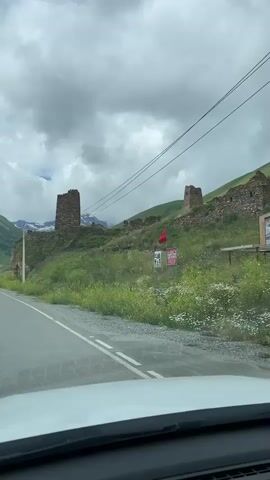 A car is driving on a paved road. On the right side of the road, there are two stone towers on a green hill. There is a red flag and signs in front of the towers. The sky is cloudy.