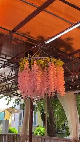 A pink floral chandelier with green leaves hangs from an orange canopy. The chandelier is suspended by chains from the center of the canopy's metal frame. The canopy is orange and appears to be made of fabric or plastic, with a metal support structure visible. In the background, through the canopy's edges, trees with green foliage and a light-colored building can be seen. White sheer curtains are also visible hanging on the right side.