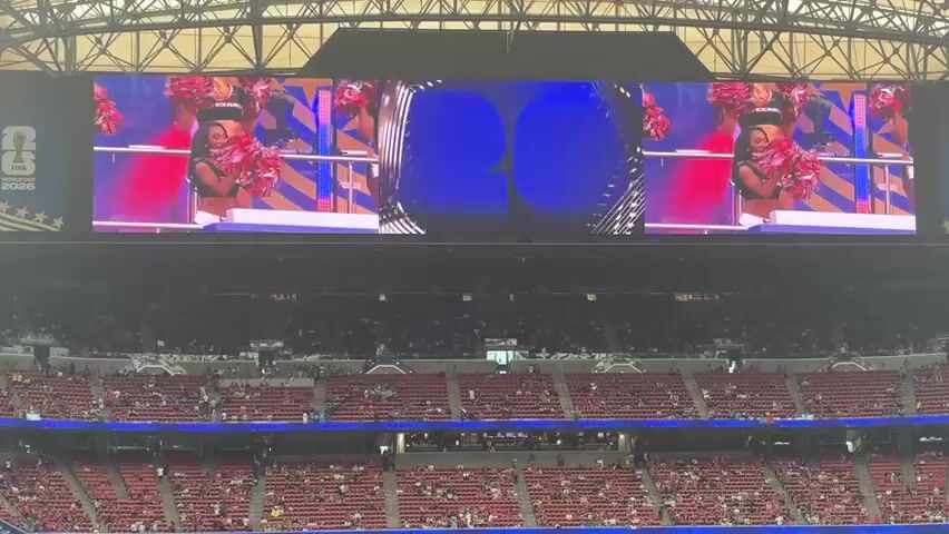 This media file captures a wide shot of a stadium interior. The main focus is a large, multi-panel LED screen displaying a variety of content. In the left and right panels of the screen, two different groups of cheerleaders are shown. The cheerleaders are wearing athletic attire, some in red and black, with "TEXANS" visible on some of their outfits. They are posing with pompoms. The central panel of the screen features abstract graphics, including what appears to be a trophy or award, and a blue background with stars.

Beneath the screen, tiered seating filled with spectators is visible, indicating a live event. The stadium appears to be indoors, with a complex metal truss structure forming the ceiling. The lighting suggests it is daytime, and the atmosphere seems energetic, likely during a sporting event or pre-game show given the presence of cheerleaders and the stadium setting. The visible text on the screen includes "TEXANS" and "FIFA WORLD CUP 2026", suggesting the event is related to soccer and possibly a celebratory announcement or promotion. The overall impression is one of a large-scale public gathering and entertainment event.