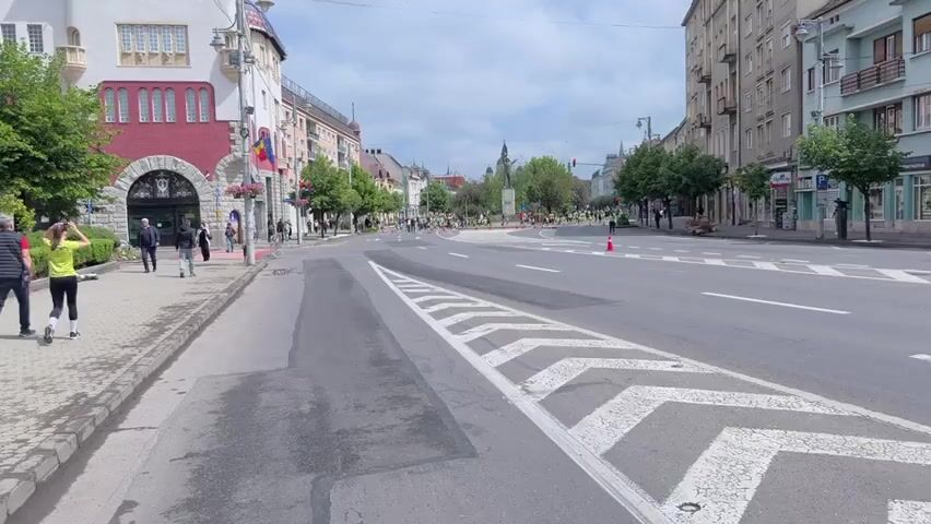 The video shows a wide, paved street in a European city, likely Romania based on the flags seen on a building to the left. In the distance, a large group of people are walking, many wearing bright yellow or green shirts, seemingly participating in a coordinated event. They are moving around a roundabout area, marked by red traffic cones.  The roundabout contains a park with a statue at its center.  The street itself has white painted lane markers and a section of chevron patterns.  Several pedestrians stroll along the sidewalk to the left, and buildings line both sides of the street, including shops and apartments. The sky is overcast with white clouds.  The overall impression is a somewhat calm city scene with a planned pedestrian event taking place.