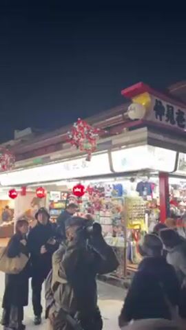The video shows a crowded, brightly lit street at night, possibly in Japan. Many people are walking down the street, some wearing traditional Japanese clothing like kimonos. There are shops lining both sides of the street with awnings decorated with strings of red and white decorations. Overhead, similar decorations hang across the street, creating a festive atmosphere. The overall impression is a bustling and vibrant scene.