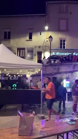 A nighttime outdoor gathering is depicted. A white tent structure is positioned on the left, with a sign visible that reads "L'Orangerie Café". Several people are standing and milling about. In the foreground, a woman with dark hair and a light-colored jacket is looking forward with her hands clasped. To her left, a woman with glasses and a gray shirt is also looking forward. Behind them, various individuals are visible, some interacting and some standing. Buildings with lit windows form the backdrop. The scene appears to be a casual event, possibly a street festival or outdoor cafe.