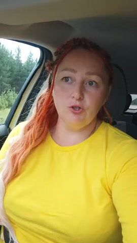 A woman with bright orange and blonde braided hair, wearing a yellow t-shirt, is looking at the camera from inside a car. She appears to be speaking. The background outside the car window shows trees and greenery.