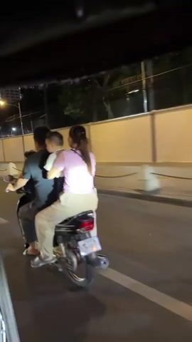 A nighttime video shows a family of three riding on a motorcycle. A man is driving, a woman is in the middle, and a child is on her lap. The motorcycle is black and appears to be in motion. The background is dimly lit, with a light-colored wall and some trees visible. The overall impression is of a common scene of family transportation.