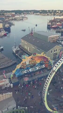 The video shows an aerial view of a waterfront carnival. The "Seven Seas" ride, featuring a pirate ship theme, is prominently displayed. A large Ferris wheel is also visible, with its lights illuminated. Crowds of people are gathered on the boardwalk, enjoying the carnival atmosphere. Boats are docked in the harbor, and buildings line the waterfront. The sky is overcast, suggesting either early morning or late evening.