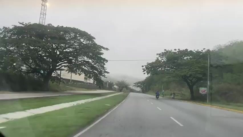 The video displays a first-person perspective from a vehicle moving forward on a wet, two-lane asphalt road under an overcast and misty sky. A grassy median with trees and shrubs separates the road from a large, modern, cream-colored building featuring large windows and a curved facade, situated on the left. A palm tree is visible in front of the building. On the right side of the road, a lush green hillside slopes upwards, adorned with trees and utility poles. Several individuals are standing on the shoulder of the road on the right. A single motorcycle travels ahead in the same direction as the camera vehicle. Distant hills are partially obscured by fog or mist.