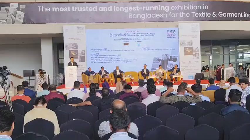 The video shows an indoor event, likely a seminar or conference, with a seated audience facing a stage. A large banner hangs across the top of the background, reading "The most trusted and longest-running exhibition in Bangladesh for the Textile & Garment industry." On the stage, a panel of speakers sits behind a table, facing the audience. To the left of the stage, a presenter stands at a podium. The audience members are mostly seated in rows of chairs. The environment appears to be a large exhibition hall or convention center, with an escalator visible in the background. People are present both on the stage and in the audience, with some individuals standing and moving around in the background. The lighting is bright and consistent with an indoor event space.