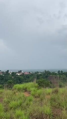 The video shows a landscape view of a village or town situated on a hillside. The foreground is dominated by tall, green grass and some reddish-brown soil. In the middle ground, there are trees and various buildings with red and brown roofs, indicating a settlement. The background features a hazy horizon under a cloudy, gray sky. The overall impression is one of an overcast day, possibly before or during a rain shower, as indicated by the narration mentioning rain. There are no people or specific actions clearly visible in the video.