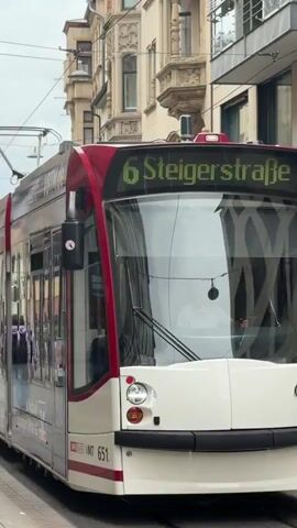 A tram is seen moving along a street. The tram is red and white and has the number 6 displayed on its front. The windows of the tram are reflective, showing the surroundings and people walking on the sidewalk. People are walking on the sidewalk, and buildings line the street. The sky is overcast. The tram is moving from left to right in the frame.