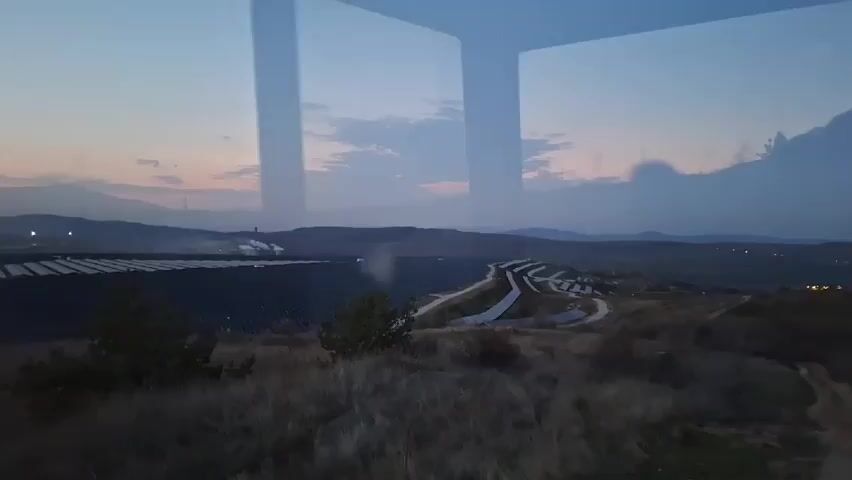 The video shows a landscape at dusk, viewed through a window.  In the distance, there's a field of solar panels and what appears to be a serpentine road with light-colored paths on either side. The landscape is hilly, with distant mountains visible on the horizon. The sky is a mix of blue and hints of orange from the setting sun, with clouds partially obscuring the view.  In the foreground, there's dry, scrubby vegetation. The window's reflection is visible, adding another layer of the outside scenery. There's a dark, rod-like object partially obscuring the view from the right.