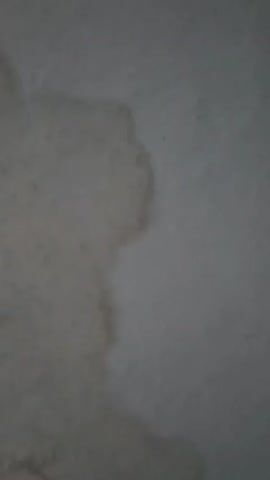 This is a close-up, shaky video of a textured surface, likely a ceiling, with a water stain. The stain is irregular in shape and has a yellowish-brown hue, indicating it is likely old. The lighting is dim, creating shadows and making it difficult to discern fine details. The video pans slowly across the stain, showing its extent and variation in color. There are no people or other objects visible in the frame. The environment appears to be indoors.