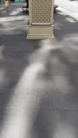A video opens on a paving stone walkway with dappled sunlight and shadows. A tall, decorative clock tower with an ornate pattern on its base stands in the center of the frame, slightly off to the right. A young child wearing a yellow dress walks away from the clock tower on the left side of the frame, holding onto a hula hoop. A woman in black clothing walks behind the child. In the background, there are several multi-story buildings with windows and balconies, as well as a bridge and what appears to be a pool or water feature. A few benches are visible along the walkway. The camera pans slightly to the right, revealing storefronts on the right side of the frame.
