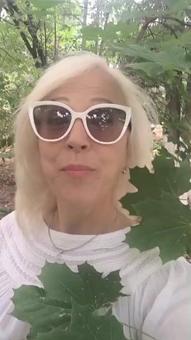A blonde woman wearing white sunglasses and a white ruffled shirt smiles at the camera. She is outdoors in a wooded area, with green leaves in the foreground. She appears to be speaking to the camera.
