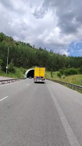 The video shows a yellow truck driving down a highway towards a tunnel. The truck is in the center of the frame, and the tunnel is visible in the distance. The highway is lined with trees and grassy hills. The sky is cloudy.
