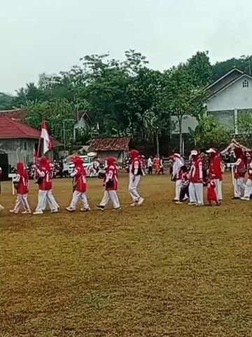 A group of children wearing red and white uniforms are walking across a grassy field. They appear to be participating in a parade or a performance. The children are accompanied by adults, some of whom are also wearing similar uniforms. In the background, there are houses, trees, and a cloudy sky. The overall atmosphere appears to be festive and celebratory.