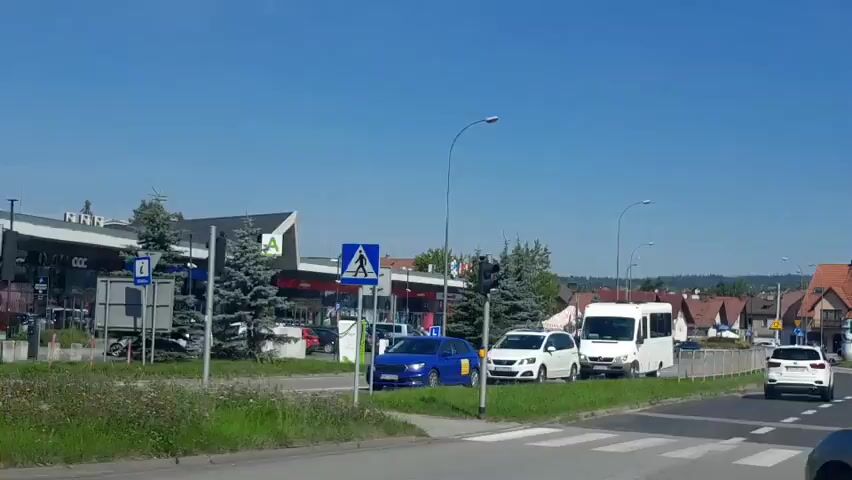 A bright, sunny day with a clear blue sky is depicted. The scene is a street view of a shopping complex. In the foreground, there is grass and some plants. To the left, a street light pole and a traffic signal are visible. A person on a bicycle rides past on the sidewalk. The shopping complex features multiple storefronts with signs in Polish. Visible brands include "CCC," "KARA," and "Netto." There are several cars parked in front of the stores and some driving on the street. A pedestrian crossing signal is also visible. Trees are planted along the sidewalk in front of the complex. The overall impression is that of a typical suburban commercial area on a pleasant day.