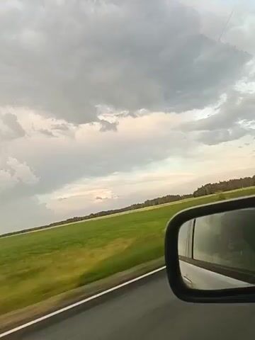 The video captures a view from inside a moving car, looking out the side window and into the side-view mirror. The exterior scene consists of a road running alongside a vibrant green field that stretches towards a treeline in the distance. The sky above is filled with dramatic, grey clouds, suggesting impending weather changes. The side-view mirror reflects a blurred image of the car's interior, possibly showing a person's head and shoulders. The motion of the car creates a slight blur in the landscape, emphasizing the sense of travel.