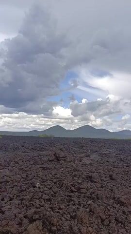 The video depicts a broad, flat landscape composed of dark, rocky terrain, likely cooled lava. In the distance, a range of hills is visible, with a strip of green vegetation along the horizon, separating the rocky ground from the hills. The sky is filled with a mixture of gray and white clouds, partially obscuring a patch of blue sky. The camera pans slightly, offering a wider view of the expanse of lava rock and the sky above.