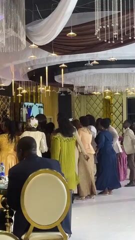 The video shows an indoor event hall with elegant decor. Suspended from the ceiling are draped white fabric, brown fabric, and crystal chandeliers. Several small, circular light fixtures hang from the ceiling as well. In the center of the frame, a group of people, predominantly women dressed in formal attire, are dancing and interacting. A man in a dark suit is seated in the foreground, his back to the camera, at a table with a gold-rimmed chair. Another man in a light-colored shirt and dark pants stands further back in the group. In the background, a large screen displays an image of several people standing. The floor appears to be white. The overall atmosphere suggests a celebratory occasion like a wedding or formal party.