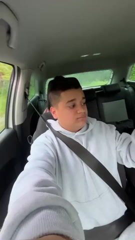 A young person wearing a light gray hoodie sings and drinks from a can in the passenger seat of a moving car.  Someone in the back also drinks from a can.  The camera angle shifts to the front of the car showing the view of a paved road lined with trees. The sky is partly cloudy.
