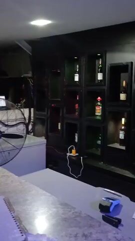 The video shows the interior of what appears to be a bar or nightclub. In the foreground, a bar counter is visible with a spiral-bound notebook on it. Behind the counter, a large industrial fan is positioned next to a white refrigerator. Further back, a dark shelving unit holds several bottles of alcohol. A person in a blue patterned shirt walks across the background. The lighting is dim and atmospheric, with blue and white lights illuminating the space. There is a shelf with bottles in the background that is lit up, and a person is seen in the background behind the bar. The fan is turned on and blowing air. The overall scene is dimly lit and appears to be a service area of a bar. The refrigerator is white and appears to be a commercial size. The bottles on the shelf are of various shapes and sizes, some with labels visible. The person in the blue shirt is blurred due to motion. The fan is a large, metal oscillating fan. The notebook on the counter has writing on it, but it is not legible. The environment appears to be indoors.