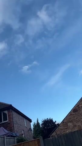 The video shows a view upwards towards a bright blue sky with scattered white clouds. In the lower part of the frame, a wooden fence runs horizontally across the bottom. Behind the fence, there is a brick house with a dark roof and a window. Further in the background, there are several trees with dark green foliage. The scene appears to be outdoors during daylight.