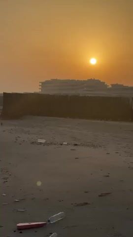 A beach at sunset. The sky is orange. A sun is in the sky. There are buildings in the background. There is a fence made of reeds in the foreground. There is trash on the beach, including plastic bottles and a pink object.