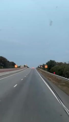 A first-person perspective video captures a view from a vehicle driving on an empty highway. The road ahead is wet and appears to be a multi-lane highway with a dividing barrier on the left. There are speed limit signs visible on both sides of the road, indicating 70. The sky is overcast and a pale blue-gray. Trees and foliage line the sides of the highway. There are a few distant vehicles visible on the road, and their headlights are on. The overall impression is that of driving on a highway in overcast weather, possibly in the early morning or late evening.