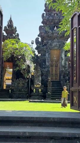 The video presents a medium, wide shot of a courtyard of a Balinese temple with a young boy standing on the grassy area in the center. The boy is facing toward the camera wearing a traditional Balinese outfit of a light green shirt and a brown and gold skirt. Two ornate, dark stone Balinese temple structures are to the left and right of the boy. Each structure has steps leading up to an entrance. The shot is framed by an open dark brown wooden gate in the foreground. The weather is sunny and the sky is blue. There is also a sound of traditional Balinese music playing.