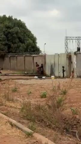 The video shows a roadside scene where people are washing clothes. In the background is a tall wall topped with barbed wire. There's a large green tree on the left. Three people are near a tap washing items. Two other individuals are squatting or bent over, seemingly washing clothes on the ground. The environment is dry and the ground is mostly reddish-brown with sparse vegetation. There are some buckets and other items near the people. A metal tower stands in the distance to the right. The sky is overcast.