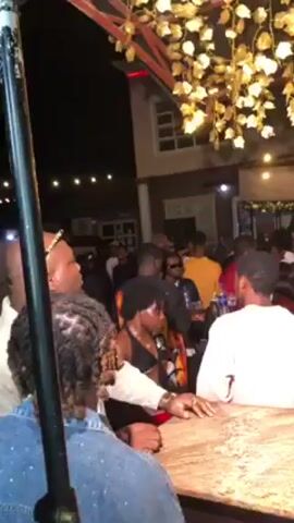 The video shows an outdoor party at night. Numerous people are visible, mostly standing, talking, and moving to the music. Some individuals are near a bar. Overhead, there is decorative lighting resembling flowers. Buildings are visible in the background.