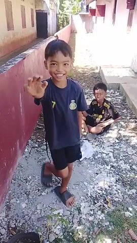A young boy with dark hair and brown skin, wearing a dark t-shirt with a yellow number 9 and the word "MBAPPE" on the back, is shown. He is standing and raising his right hand towards the camera, palm facing forward, as if to say hello or stop. He is smiling. The background appears to be an outdoor alleyway or path between two buildings, with a red wall on the left and a white building on the right. The ground is covered with gravel and debris. In a subsequent frame, the boy is seen sitting on the ground, drinking from a bottle, and then walking away from the camera, facing towards the white building.