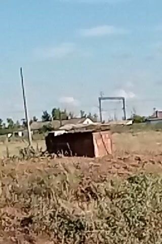 The video shows an outdoor scene with a small, weathered, rectangular structure in the foreground. Behind it, there is a flat, dry grassy field with scattered bushes. In the middle ground, a white vehicle is visible, appearing to be a van or SUV. The vehicle is stationary with its rear doors open. Further in the background, there are several buildings, some appearing to be houses with pitched roofs, and other structures that could be outbuildings or sheds. A large metal frame structure, possibly for power lines or construction, is also visible in the distance. The sky is clear and blue with a few wispy clouds. The lighting suggests it is daytime. The overall environment appears to be rural or semi-rural.