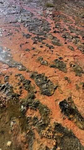 A video shows a flowing body of water with a reddish-orange hue covering the ground, likely due to mineral deposits. The water appears to be warm and steamy as it cascades down from a structure in the background. This structure seems to be a fountain or a part of a water feature, with water gushing out of a cylindrical pipe. In the distance, lush green mountains rise majestically under a clear blue sky dotted with white clouds. The scene conveys a natural, possibly geothermal, landscape.