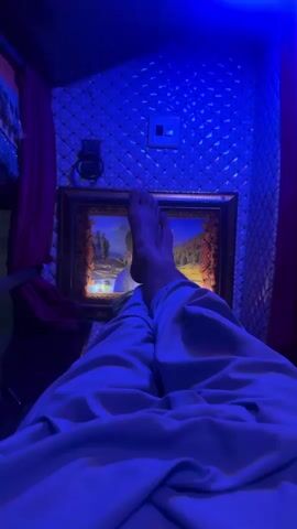 The video begins with a first-person perspective of someone relaxing, with their legs extended and covered by a light-colored blanket. A framed picture is visible on the wall behind them, and blue lights illuminate the space. The camera then pans to the right, showing a window with a blurred view of the outside. A motorcycle can be seen passing by in the distance. The interior of the space appears to be decorated with tassels and other ornamental items. The blue lighting continues throughout the clip.