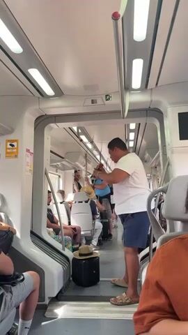 The video shows the interior of a train carriage. A man wearing a white t-shirt and blue shorts stands in the aisle, holding a hat and a phone. He appears to be talking or singing into the phone, possibly recording. Other passengers are seated on the train, some looking towards him, others looking away or at their phones. The train carriage has rows of seats, overhead luggage racks, and fluorescent lighting. There is a black suitcase with a straw hat on top near the man's feet. The environment appears to be daytime, with natural light entering from windows, which are not clearly visible. The overall atmosphere seems relaxed, with the man's actions capturing the attention of some of the other passengers.