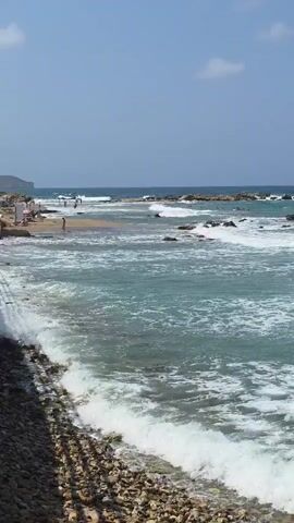 The video shows a natural beach scene. The water is turquoise with white foam from waves breaking on the shore and rocks. The sky is clear blue with a few white clouds. In the distance, there are a few people on the beach and rocks in the water. The foreground shows a stony beach and the edge of the water.