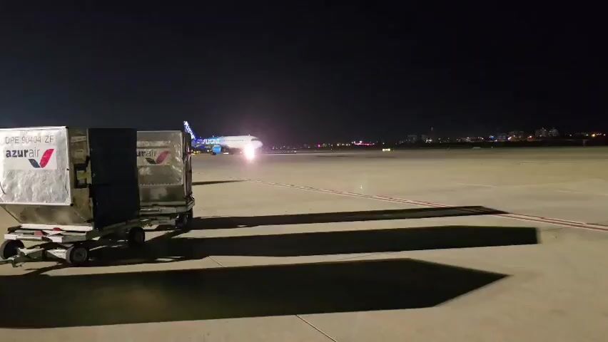 At night on an airport tarmac, an airplane with "FLYONE" visible on its side is taxiing. The plane is white with blue and colorful designs. Its landing lights are on and illuminate the runway. In the foreground, two cargo dollies are parked. The dollies have silver containers on them, one of which has the "Azur Air" logo. The sky is dark, and city lights are visible in the distance. The ground is paved, and runway markings are visible.