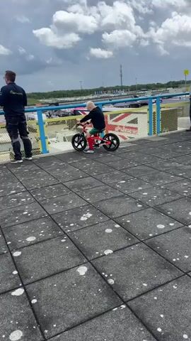 A child is sitting on a balance bike, facing away from the camera. The child has blonde hair and is wearing a black long-sleeved shirt, green shorts, and red socks. The balance bike is red and black. The child is positioned on a grey tiled surface with white spots. A blue and yellow fence is behind the child, and beyond the fence is a race track with cars and flags. A person in a black shirt and dark pants stands to the left of the child, also facing away from the camera. The sky is cloudy.