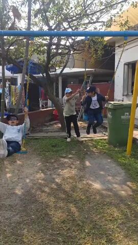 The video shows three young people playing on swings in what appears to be a backyard or park setting. The swing set is made of blue and yellow metal with chain supports. Two of the young people are swinging, while the third stands nearby, seemingly observing or waiting for a turn. All three are wearing casual clothing and baseball caps. The ground is a mix of grass and dirt. In the background, there's a white building, a tree, and some miscellaneous objects. The weather appears to be sunny.