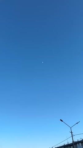 The video shows a clear, blue sky with a crescent moon visible. There are also several kites flying, one of which appears to be quite small and farther away than the others. In the lower part of the frame are utility poles and the top of a building, suggesting the video was taken in an urban environment. The overall feel is bright and peaceful, capturing the contrast of man-made and natural elements in the sky.