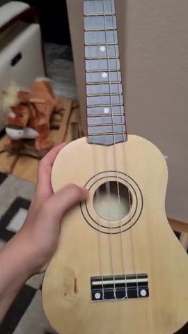 The video shows a close-up, overhead shot of a person's hand holding a light-colored ukulele. The hand is positioned over the soundhole, and it appears to be strumming or touching the strings. The ukulele is made of wood and has four strings. The background is slightly blurred, showing what appears to be a room with a rug on the floor and some objects in the distance, including what looks like a toy horse. The lighting is natural and appears to be coming from above. The video captures a brief moment of interaction with the ukulele.