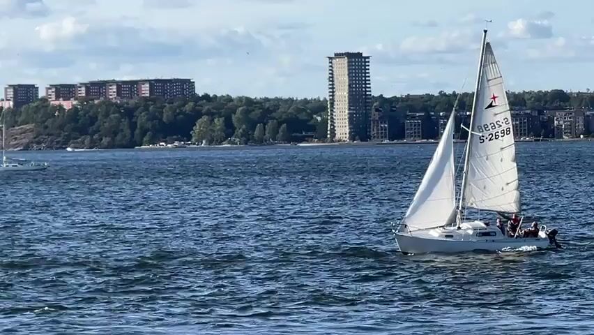 The video shows a bright, sunny day with a slightly cloudy sky over a body of water. In the foreground, a white sailboat with its sails fully deployed is sailing to the right of the frame. There are at least three people visible on the deck of the sailboat. In the middle ground, another sailboat with its sails down is visible further in the distance. In the background, a city skyline with several tall buildings and apartment complexes lines the shore, which is covered in trees. The water appears choppy, indicating a gentle breeze. The overall scene is peaceful and depicts recreational sailing in a scenic, urban environment.