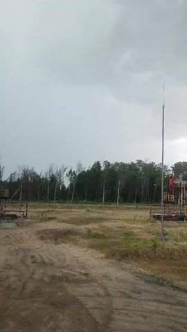 This media file captures an outdoor scene, likely an oil field or industrial area, under a cloudy sky. The prominent subjects are two visible oil pump jacks, positioned on either side of the frame. They are red and yellow mechanical structures characteristic of petroleum extraction. In the background, a dense line of trees forms a dark green border against the sky, with some sparser, bare trees interspersed, suggesting a transitional season. The ground is a mix of dirt and sparse vegetation, with tire tracks indicating vehicle movement. A tall, thin pole with an antenna-like structure stands to the right of the frame.

The activity suggested is ongoing industrial operations, with the pump jacks appearing to be in a state of readiness or operation, though their movement isn't explicitly shown in this static frame. The time of day is difficult to pinpoint precisely due to the overcast conditions, but the lighting suggests it is daytime, possibly late afternoon or early morning given the muted light. The weather appears to be cloudy and potentially unsettled, contributing to a somewhat somber or utilitarian mood.

The location context provided places this scene in Ровенскаслабодскі сельскі Савет, Belarus. There is no visible text within the frame. The overall impression is one of an industrial landscape situated near a natural environment, under a sky that hints at impending weather changes.
