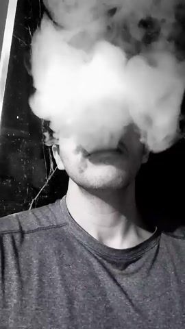 The video shows a man wearing a gray t-shirt with a small portion of his face visible. He exhales a large plume of white smoke or vapor, which quickly obscures his features. The background appears dark and unclear. The video is shot in black and white.